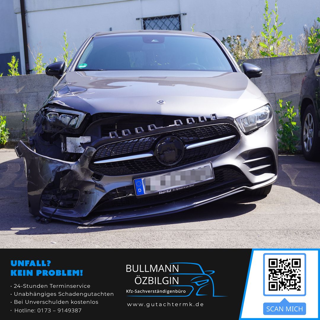 Vorderansicht eines silbernen Mercedes mit stark beschädigter linker Fahrzeugfront, fehlendem linken Scheinwerfer und verformtem Kotflügel, geparkt vor einer Mauer.