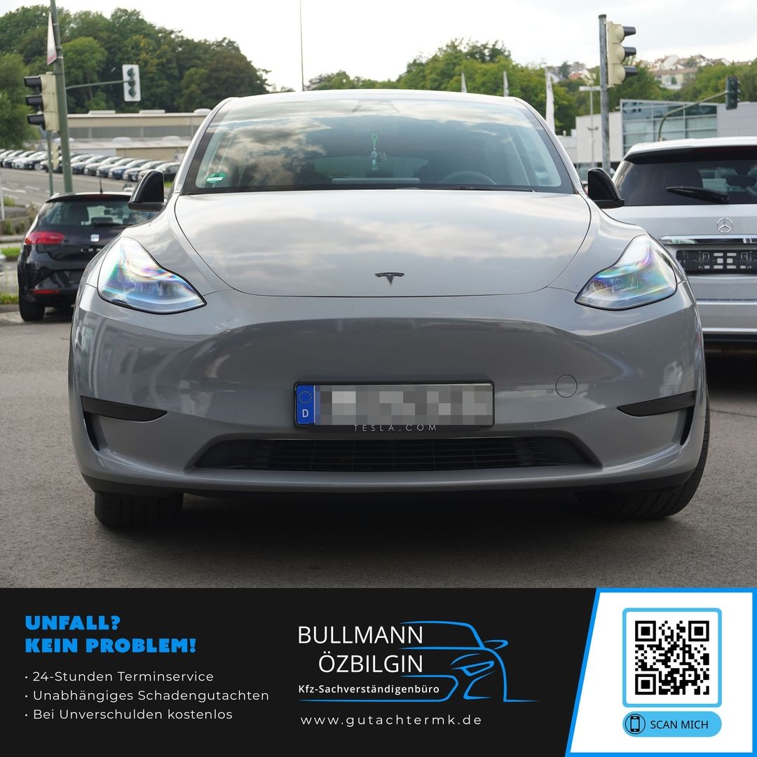 Frontansicht eines grauen Tesla Model Y auf einer Straße mit mehreren Fahrzeugen im Hintergrund.